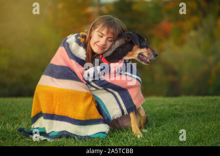 Frau eingehüllt in Wolldecken umarmen Hund im Herbst Park. Schöne glückliche junge Kaukasier Frau sitzt mit inländischen Hund Tier Haustier. Stockfoto