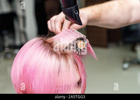 Friseur trocknet rosa Haare mit Fön und Bürste. Friseur föhnen die Haare kurz Rosa Frau hautnah. Stockfoto