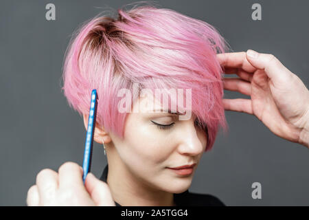 Hände von Friseur Kontrollen kurz rosa Haare des Mädchens auf grauem Hintergrund. Friseur Kontrollen mit Kammaufsatz kurz rosa Frisur der jungen Frau. Stockfoto