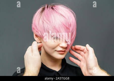 Hände von Friseur ist Kontrolle kurz rosa Haare des Mädchens auf grauem Hintergrund. Friseur Kontrollen kurz rosa Frisur der jungen Frau. Stockfoto