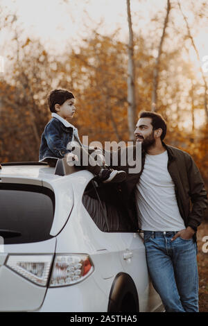 Gerne Vater stanting in der Nähe des Auto mit seinem kleinen Sohn sitzen auf dem Dach. Stockfoto