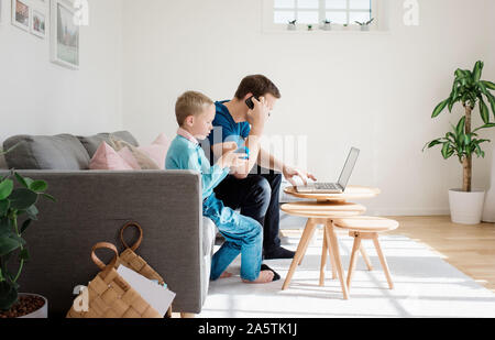 Vater saß zu Hause mit seinem Sohn auf seinem Handy und Laptop Stockfoto