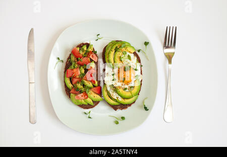 Zwei Toasts mit dunklem Brot mit Avocado, Scheiben, gehackten Tomaten, Spiegelei und microgreen. Frühstück Konzept. Ansicht von oben, weißen Hintergrund. Gesundes Essen. Stockfoto