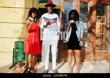 Gruppe von drei modernen trendigen afro Frankreich Freunde gegen atm aufgeworfen. Schwarzer afrikanischer Mann Modell mit zwei Dunkelhäutige womans mit Kreditkarten. Stockfoto