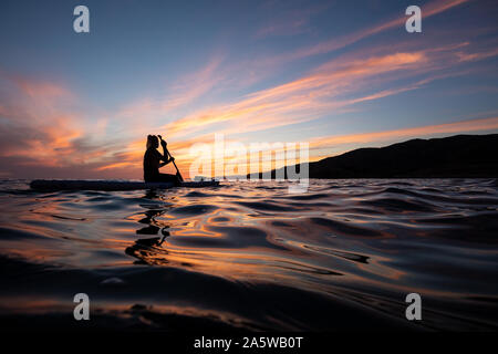 Eine Frau sitzt und Paddel auf einem wogenden Ozeans während einer lebendigen Sonnenuntergang Nachleuchten. Stockfoto