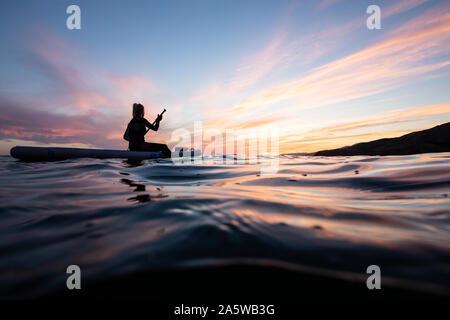 Eine Frau sitzt und Paddel auf einem wogenden Ozeans während einer lebendigen Sonnenuntergang Nachleuchten. Stockfoto