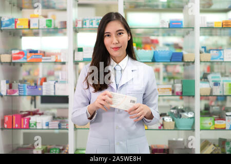 Asiatische junge Frau Apotheker mit einem reizenden Lächeln Holding medicinebox und Kamera in der Apotheke und Drogerie. Medizin, Pharmazie Stockfoto