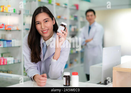 Zwei Asiatische junge Frau und Mann Apotheker mit einem reizenden Lächeln holding Medizin Flasche und Kamera in der Apotheke und Drogerie. Medicin Stockfoto