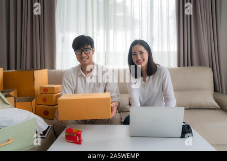 Asiatische junge Paar verkaufen on-line über einen Computer und die Verpackung der Box und Blick in die Kamera im Wohnzimmer im Haus. Small Business startu Stockfoto