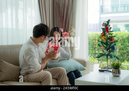 Asiatischer Mann geben die Frau roter Geschenkbox, in der es eine Flasche Parfüm. Die Frau war auf dem Sofa zu Hause abgeholt. Der Weihnachtsbaum in Th Stockfoto