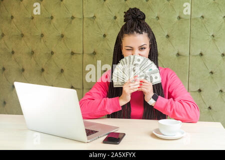 Portrait von Reichen attraktiven jungen Erwachsenen Geschäftsfrau mit schwarzen dreadlocks Frisur in rosa Bluse sitzt und zeigt viele Bargeld Dollar, coverin Stockfoto