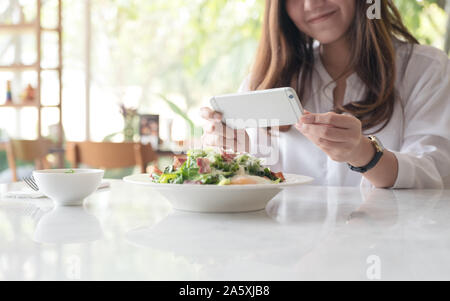 Closeup Bild eines asiatischen Frau mit Smartphone, Foto von Salat in einem weißen Teller am Tisch im Cafe Stockfoto