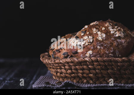 Frisch gebackene Bio Roggen Brötchen in einem voven Korb, auf einem dunklen Holztisch. Das Brot ist bestreut mit Kürbiskernen und Haferflocken. Schwarz backgr Stockfoto