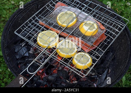 Grillen Lachs mit Zitronenscheiben auf runden Holzkohlengrill mit Rost. Heiße Glut unter Fisch. Stockfoto