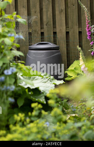 Blick auf einen schwarzen Kunststoffbehälter im Garten gegen einen Holzzaun, der durch Laub gesehen wird Stockfoto