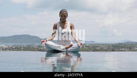 Heitere frau yoga in Lotus Haltung am Pool gegen Sky Stockfoto