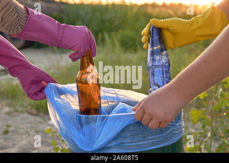 Junge Menschen in lila und gelb Gummi Handschuhe sind zu Fuß mit Müllsäcken entlang einer grünen Zone eines schmutzigen Strand des Flusses und Reinigung, Abfall, Stockfoto