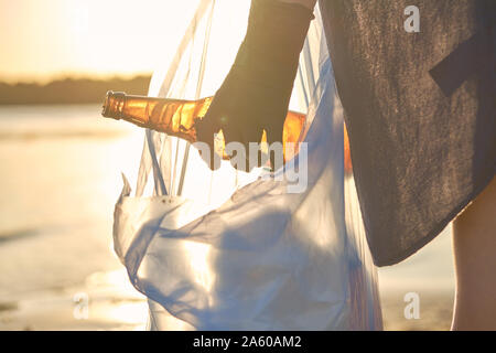 Junger Student in schwarzen Handschuhen ist Laufen mit Müllsack entlang einer schmutzigen Strand des Flusses und Reinigung sweepings, alte Glasflaschen. Menschen und Eg Stockfoto
