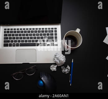 Verschiedene Objekte auf schwarz Büro Schreibtisch. Moderne schwarze Büro Schreibtisch Tisch mit Laptop Tastatur, Maus, Stift, zerknittertes Papier Kugeln, Gläser, Tasse Kaffee. Stockfoto