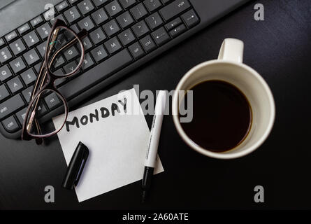 Verschiedene Objekte auf schwarz Büro Schreibtisch. Moderne schwarze Büro Schreibtisch Tisch mit Tastatur, Haftnotiz mit Montag, Gläser und Tasse Kaffee. Ansicht von oben. Stockfoto