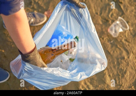 Junge verantwortliche Person in schwarzem Gummi Handschuhe ist Laufen mit Müllsack entlang einer schmutzigen Strand des Flusses und Reinigung Müll, Glas- und p Stockfoto