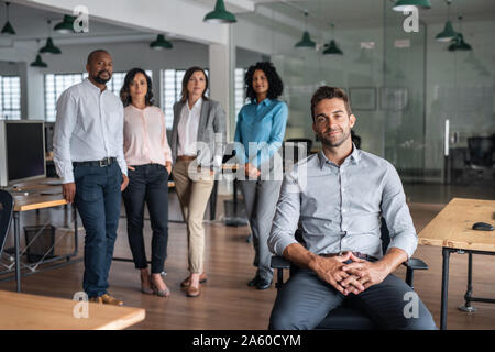 Lächelnd Geschäftsmann sitzt in einem Büro mit den Kollegen hinter ihm Stockfoto