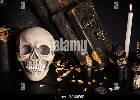 Realistisches Modell eines menschlichen Schädels mit weißen Zähne, alten Topf, brennende Kerze, Bücher mit Zaubersprüchen und Gläser trank auf Holz dunkel Tabelle sind. Die pet Stockfoto