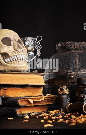 Realistisches Modell eines menschlichen Schädels mit Zähnen steht auf einem alten Bücher gegen einen dunklen Topf, Gläser und Leuchter. Die blütenblätter der getrockneten Rosen sind Scatte Stockfoto