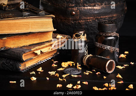 Einige Bücher mit Zaubersprüchen, alten Topf, Gläser Trank sind auf einer hölzernen dunklen Tisch. Die blütenblätter der getrocknete Rosen sind in der näheren Umgebung verstreut. Schwarzen Hintergrund. Hallowee Stockfoto