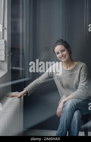 Portrait von lächelnden jungen Frau, die neben der Heizung zu Hause sitzen Stockfoto