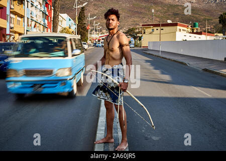 Tribal Mann mit seinem traditionellen Bogen und Pfeile in der Mitte der Stadt mit Autos vorbei, Lubango, Angola Stockfoto