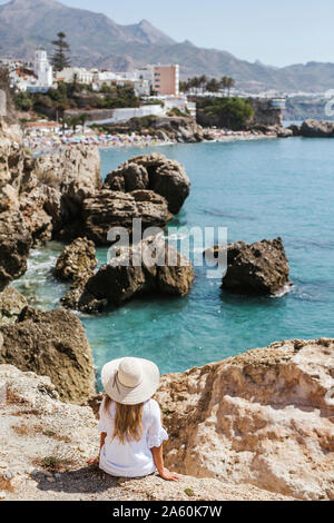 Rückansicht der Frau in weißem Kleid und Strohhut sitzen auf den Felsen auf das Meer, Nerja, Spanien Stockfoto