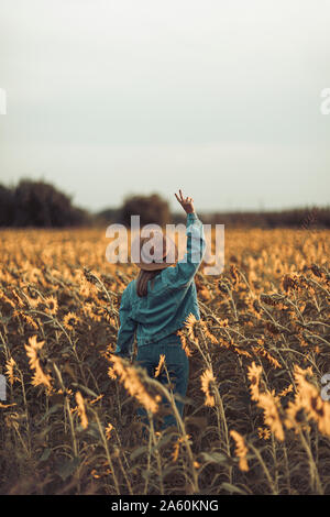 Ansicht der Rückseite des junge Frau mit Hut und Jeans Jacke in einem Sonnenblumen Feld, Frieden Zeichen Stockfoto