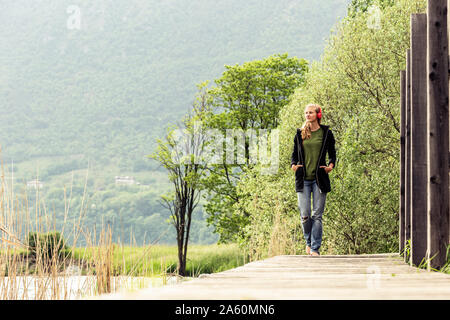 Junge Frau mit roten Kopfhörer zu Fuß auf der Promenade am See Idro, Baitoni, Italien Stockfoto