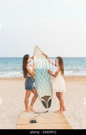 Zwei glückliche weibliche Freunde mit Surfbrett stehend auf der Promenade am Strand Stockfoto