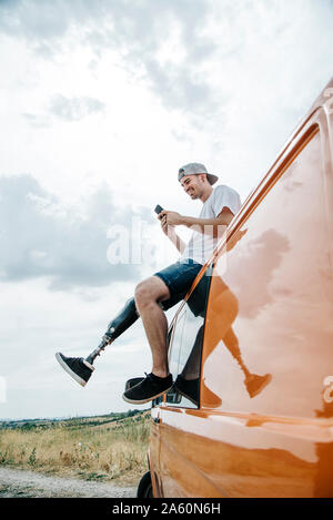 Junger Mann mit Bein Prothese sitzt auf dem Dach des Reisemobils mit Handy Stockfoto