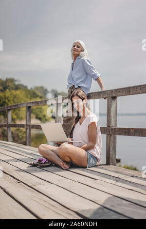 Frau sitzt auf Holz Brücke, mit Laptop, Mutter steht im Hintergrund Stockfoto