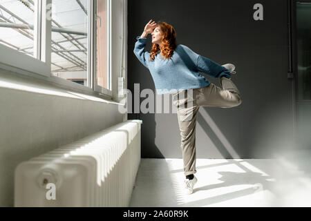 Rothaarige Frau auf einem Bein stehen in einem Loft mit Blick auf das Fenster Stockfoto