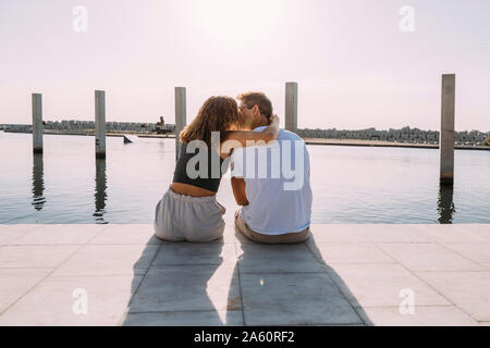 Ansicht der Rückseite des jungen Paares sitzt auf einem Pier am Meer küssen Stockfoto