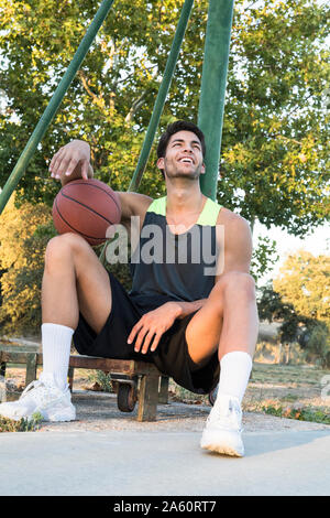 Junge Sportler sitzen auf Sportplatz holding Basketball und Lachen, wenn Sie auf der Suche Stockfoto