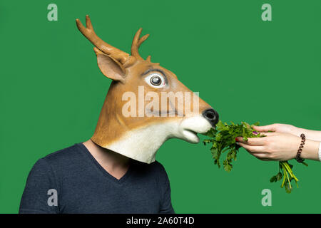 Junger Mann mit Hirsch Maske riecht Petersilie vor grünem Hintergrund Stockfoto