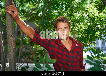 Portrait von selbstbewussten jungen Mann mit kariertem Hemd lehnte sich gegen einen Baum Stockfoto