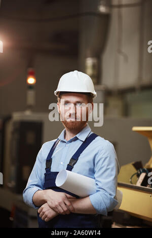 Porträt von Ernst bärtigen Manager in Overalls und in Arbeit Helm Holding Blaupause und die Kamera während der Arbeit in der Anlage Stockfoto