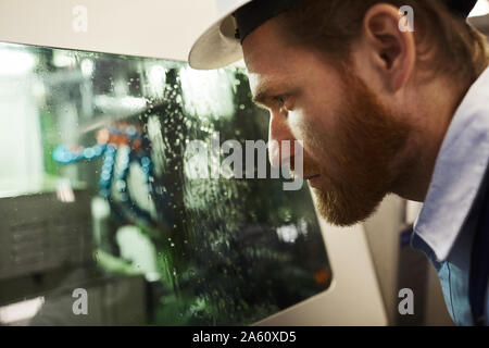 Nahaufnahme der Bärtige junge Unternehmer in Arbeit Helm durch das Glas auf der Suche und Kontrolle der Arbeit der Maschine in der Anlage Stockfoto