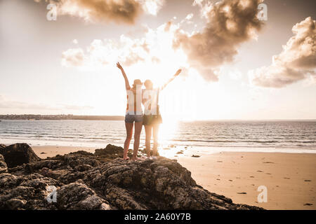 Zwei Freundinnen stehen auf felsigen Strand, winken bei Sonnenuntergang, Ansicht von hinten Stockfoto