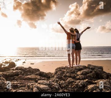 Zwei Freundinnen stehen auf felsigen Strand, winken bei Sonnenuntergang, Ansicht von hinten Stockfoto