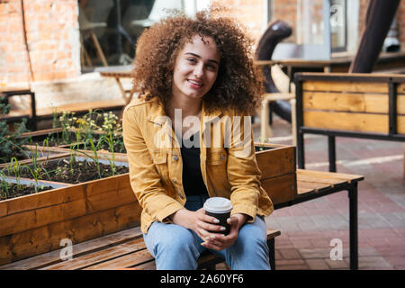 Portrait Of Smiling teenage Mädchen sitzt auf der Bank mit Kaffee zu gehen Stockfoto