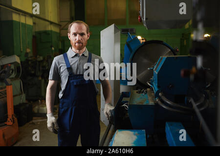 Portrait der junge Bartgeier Mechaniker in Latzhosen an Kamera suchen, während in der Fabrik stehen Stockfoto
