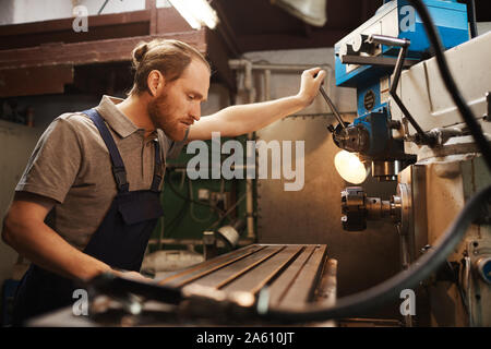 Bärtige Mechaniker in Latzhosen steuert die Maschine und für den Prozess zu beobachten, während in der Fabrik arbeiten Stockfoto