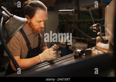 Ernsthafte bärtigen Mechaniker arbeiten mit metallischen Objekten in der Nähe der Drehmaschine in der Fabrik Stockfoto